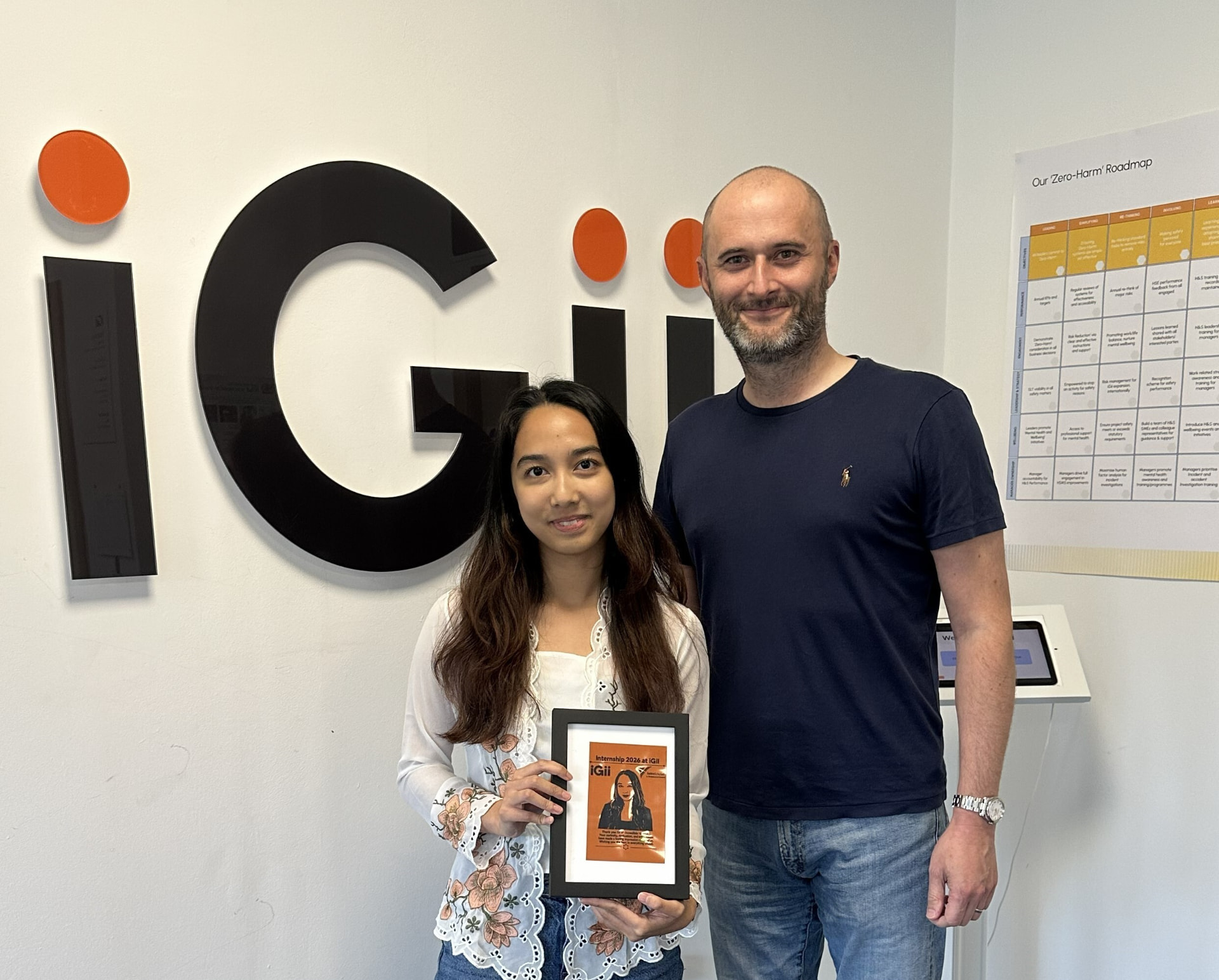 Saltire Scholar Arisya holding her framed Gii portrait beside a colleague, in front of the iGii logo at the Stirling office