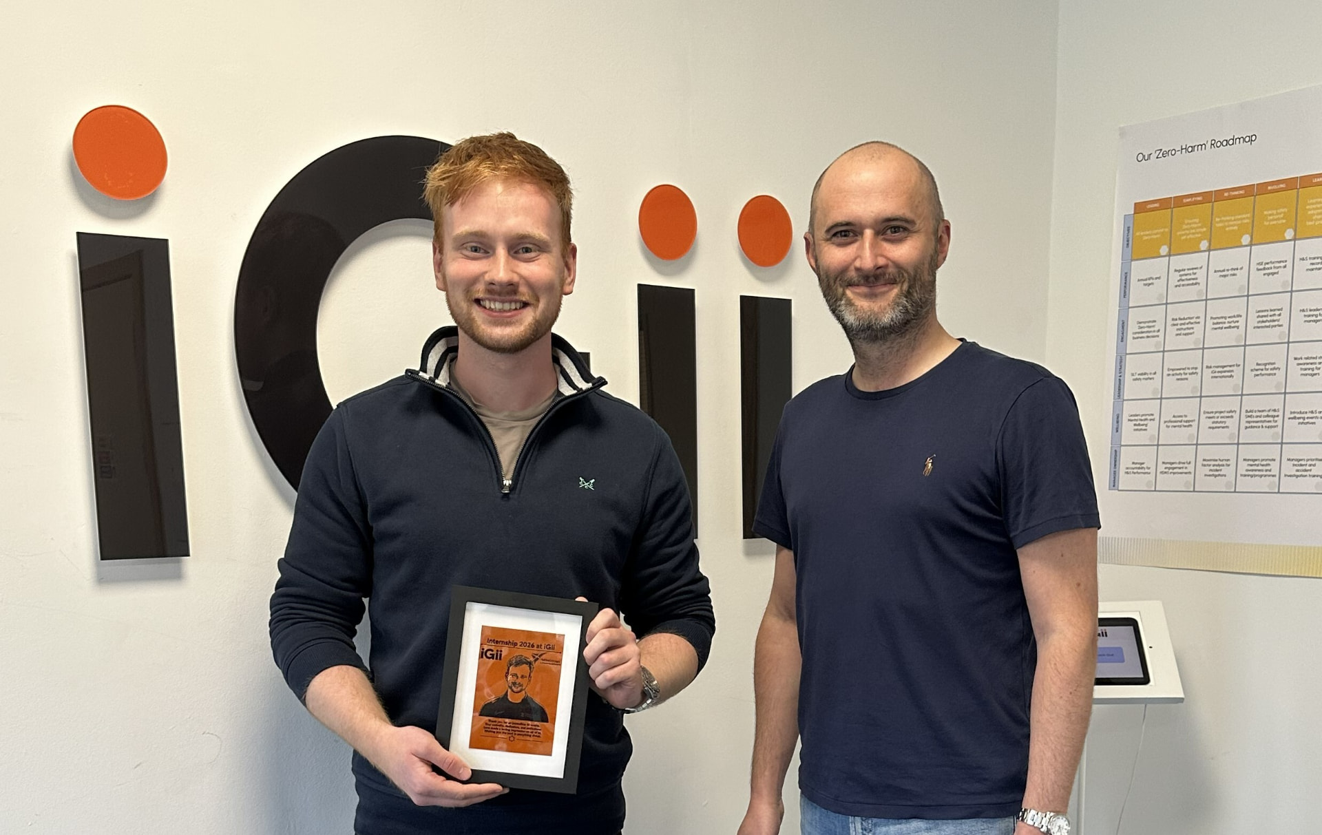 Saltire Scholar Josh holding his framed Gii portrait beside a colleague, in front of the iGii logo at the Stirling office